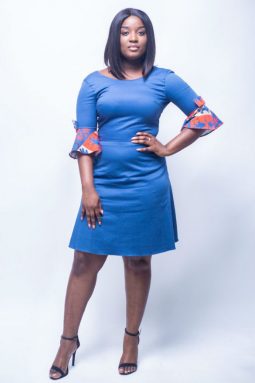Black woman looks seriously, while posing with an A-shape dress. That african inspired dress is Plain Blue. It has a sleeve with bow-tie and red decoration.