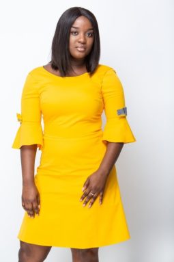 Black woman poses with an A-shape dress. It is plain yellow and has a sleeve with bow-tie.