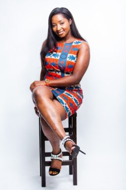 Black woman sits on a chair in a form-fitting dress called Red & Blue. It has a belt and no sleeve.